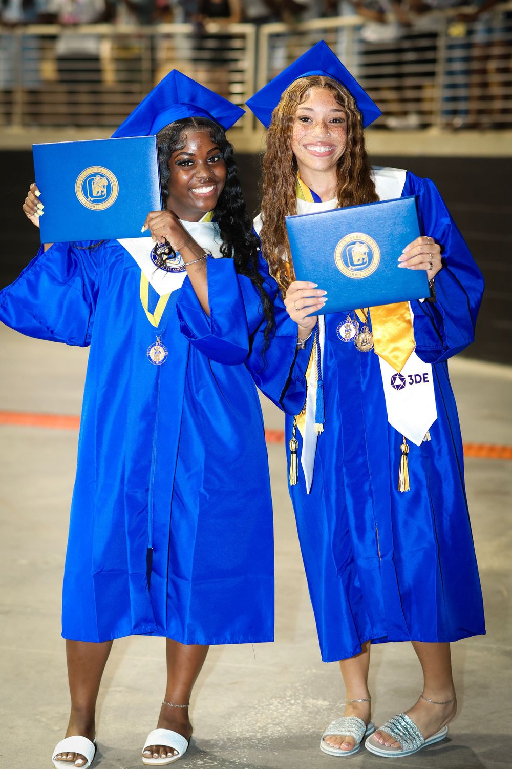 Two graduates in blue caps and gowns proudly display their diplomas at a graduation ceremony.