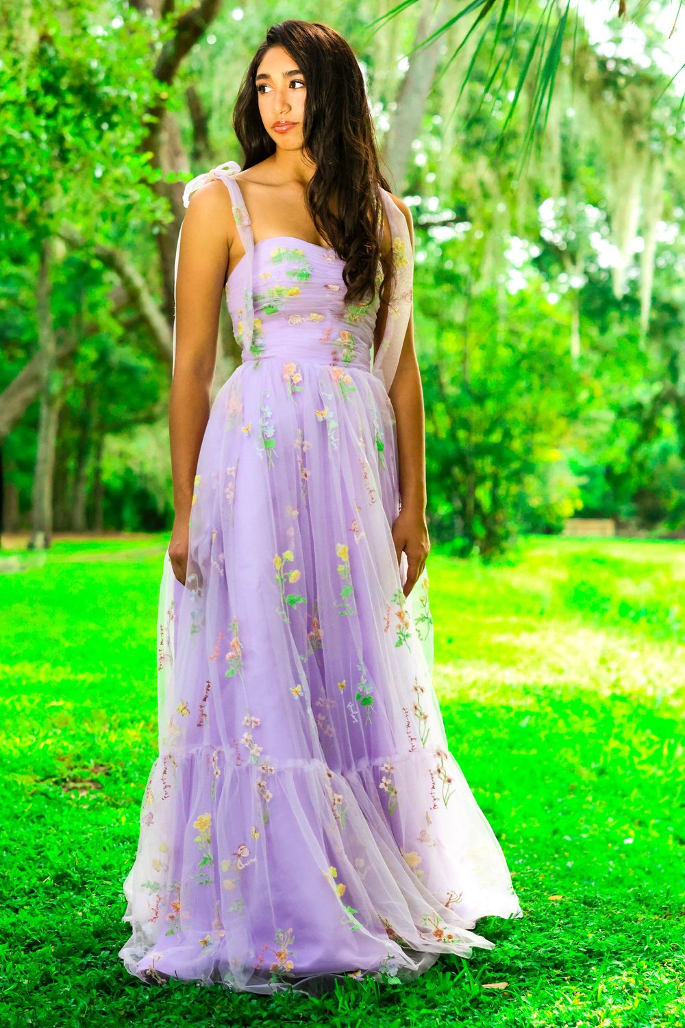 A flowing lavender tulle dress with floral embellishments is photographed against lush green garden scenery.