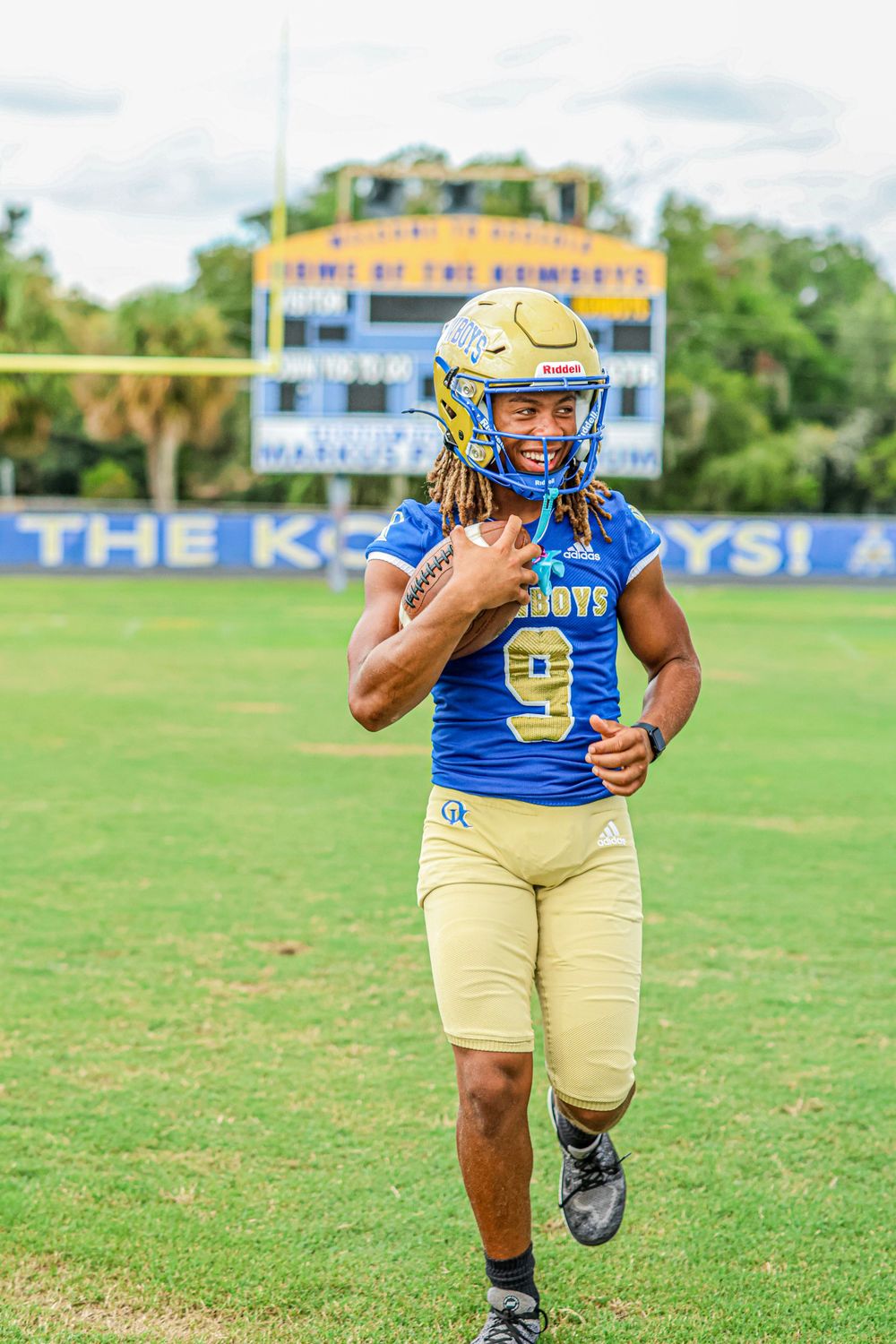 Football player in blue and gold uniform runs drills on practice field during training session.
