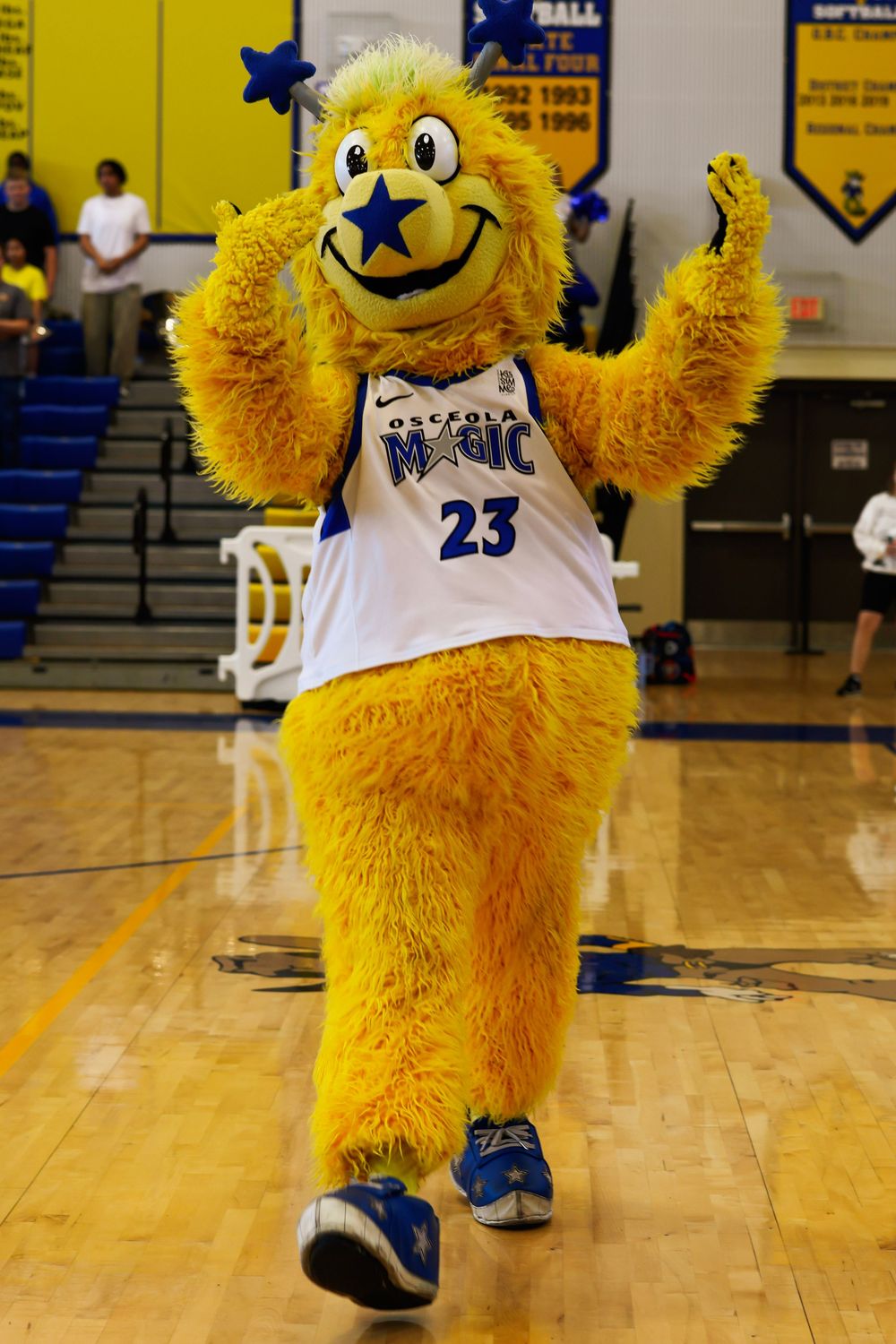 A yellow furry mascot in a white basketball jersey performs energetic dance moves on a gymnasium floor.