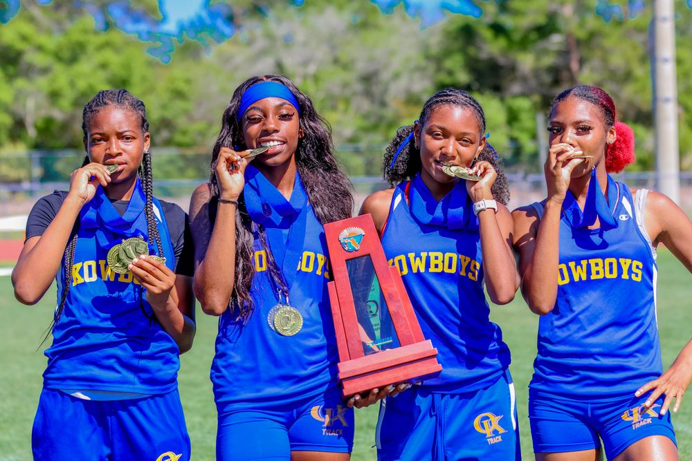 Four female athletes in blue Cowboys uniforms bite gold medals while holding a trophy on a track field.