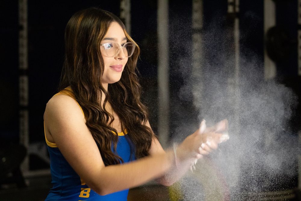 An athlete in a blue tank top applies chalk powder to hands, creating a misty cloud effect in dark lighting.