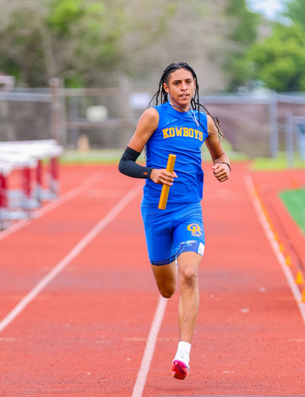 A young athlete in blue Kowboys uniform runs relay race on red track, holding baton.