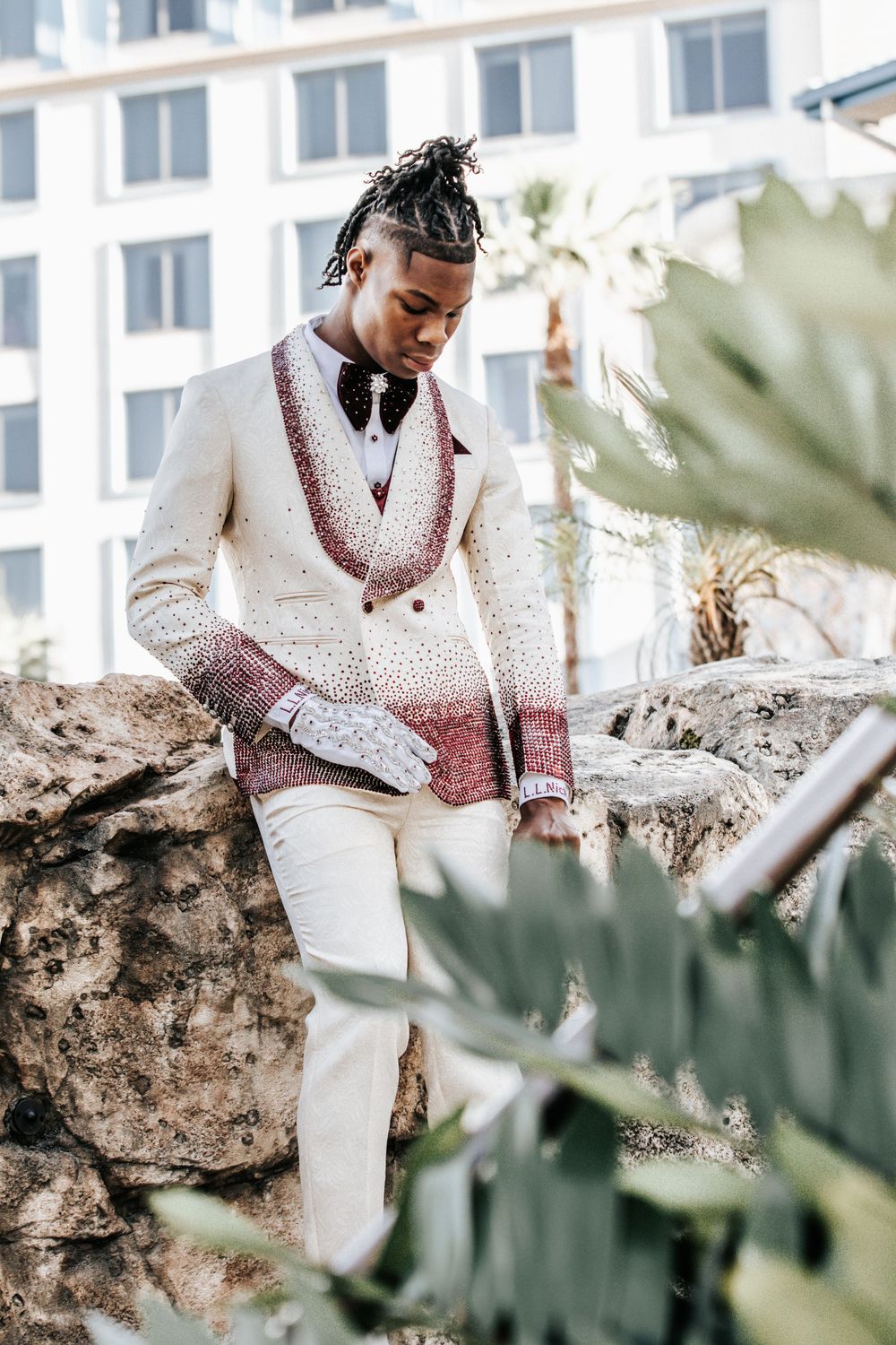 Stylish Black man in cream and burgundy embroidered jacket with white pants poses outdoors near rocks and tropical plants.