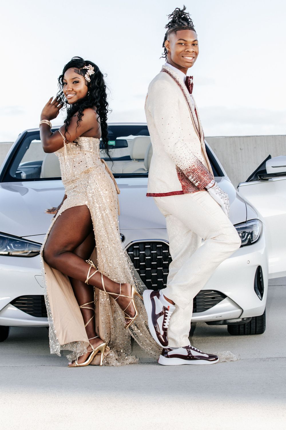 Two dressed-up teens pose in formal prom attire beside a white convertible car.