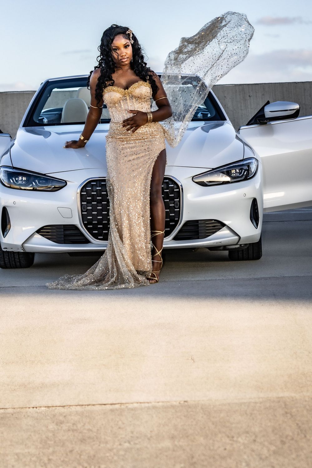A woman in a gold sequin gown poses in front of a white BMW convertible on a rooftop parking lot.