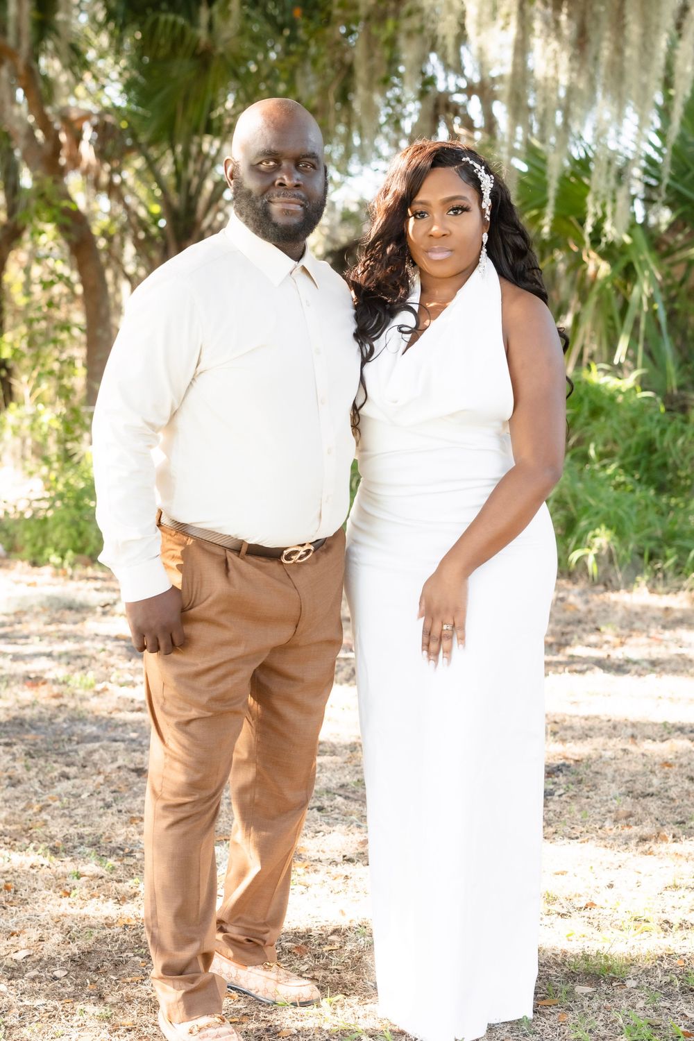 Couple posing outdoors under Spanish moss trees, man in white shirt and tan pants, woman in elegant white dress.