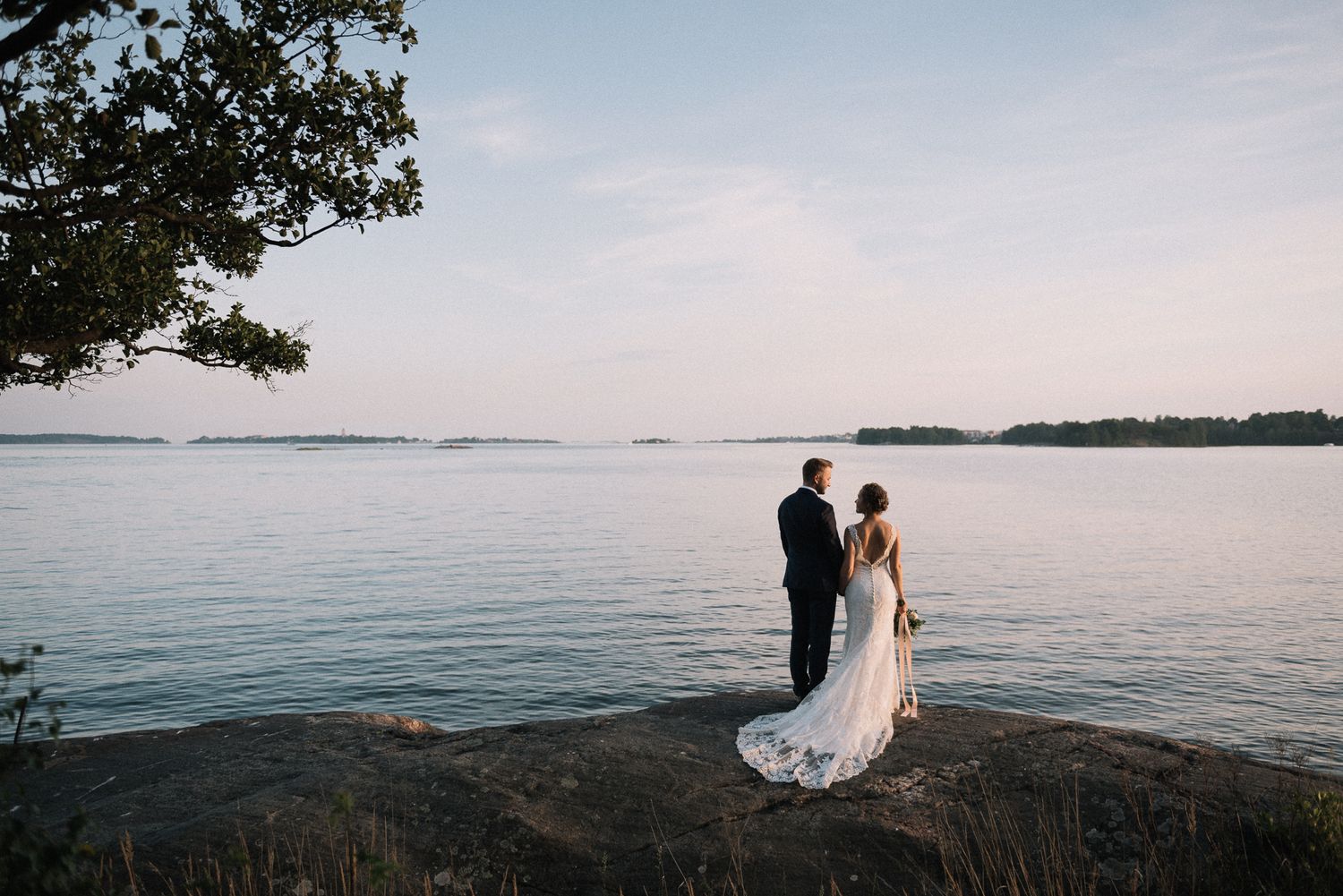 A couple stands silhouetted on a rocky shoreline overlooking a serene lake at sunset.
