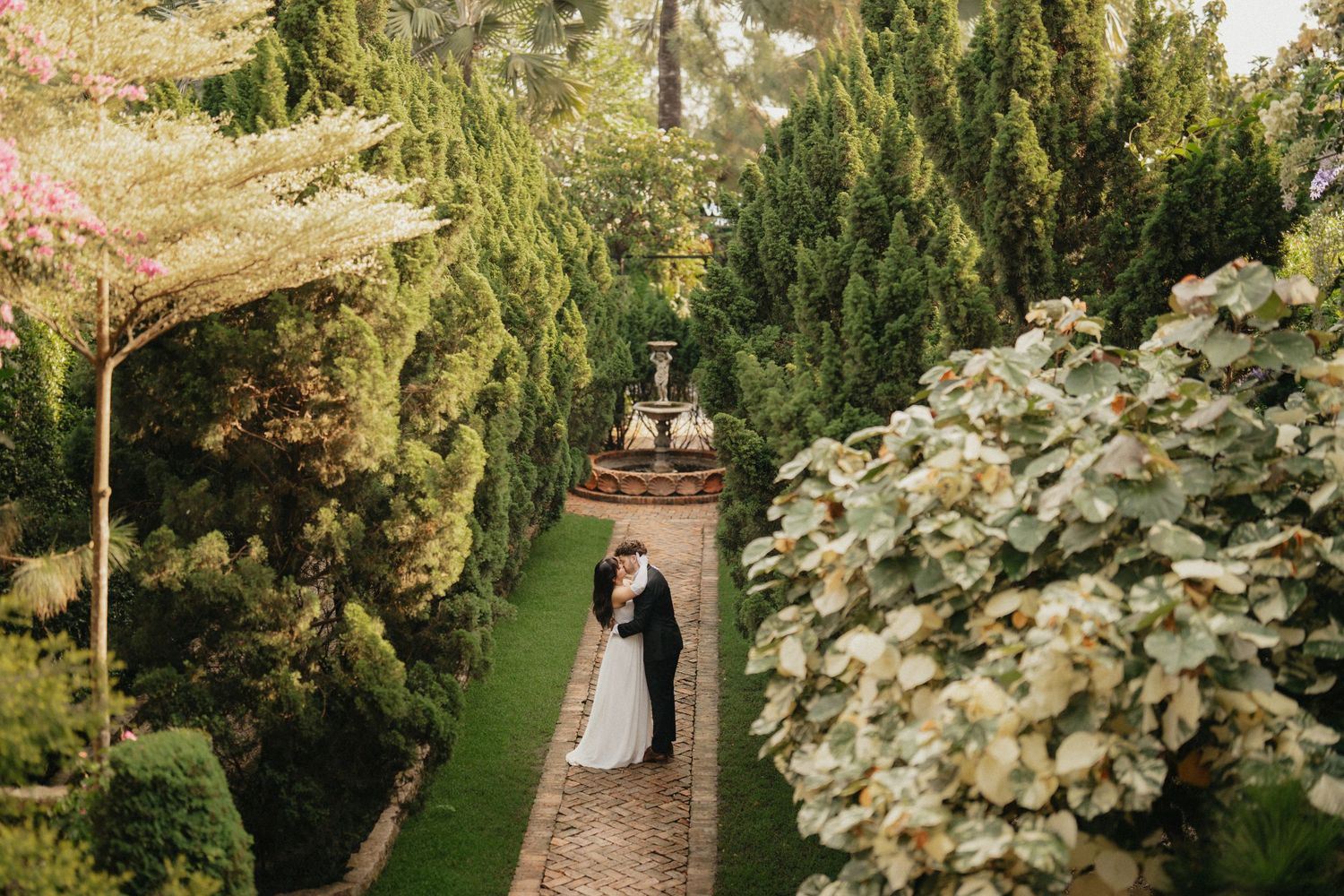 Bride and groom sharing a romantic kiss on a garden pathway surrounded by lush greenery and a decorative fountain.