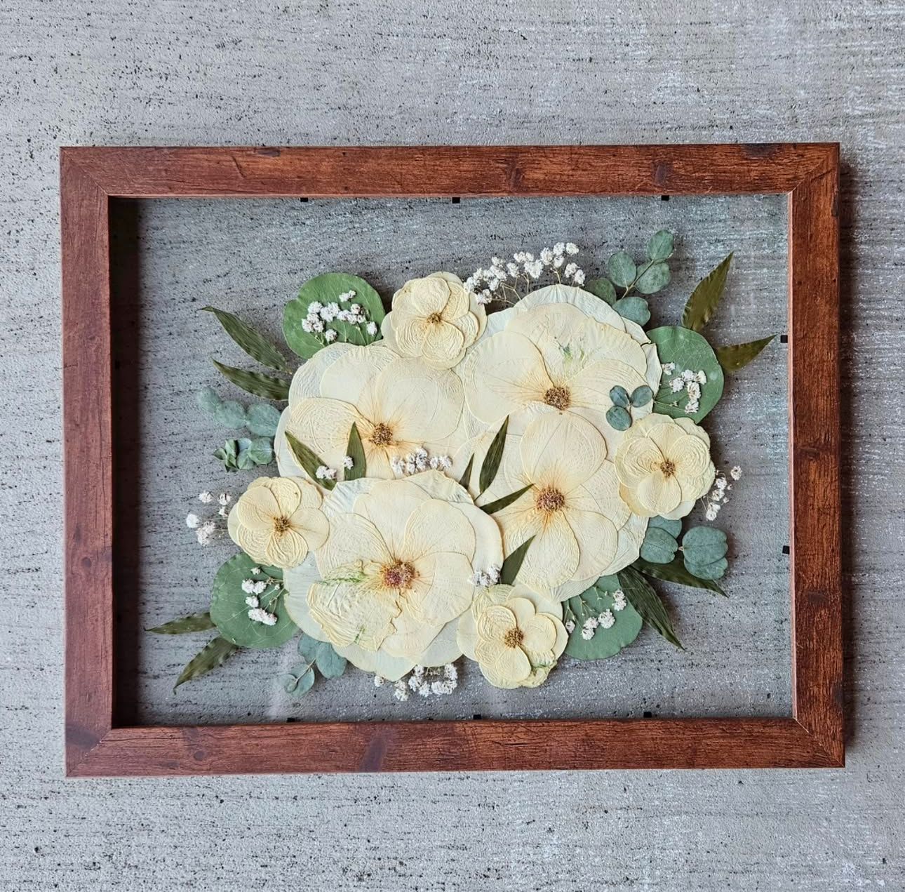 Pressed white flowers arranged in wooden frame against grey background with eucalyptus leaves and baby's breath accents.