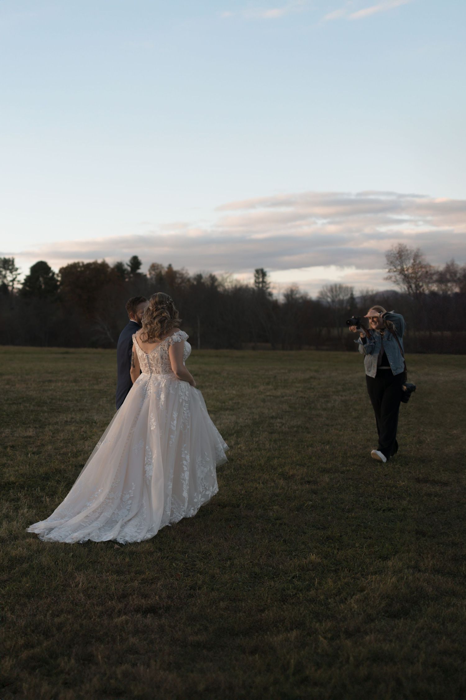 A photographer captures a bride in a flowing lace wedding dress in an open field at dusk.