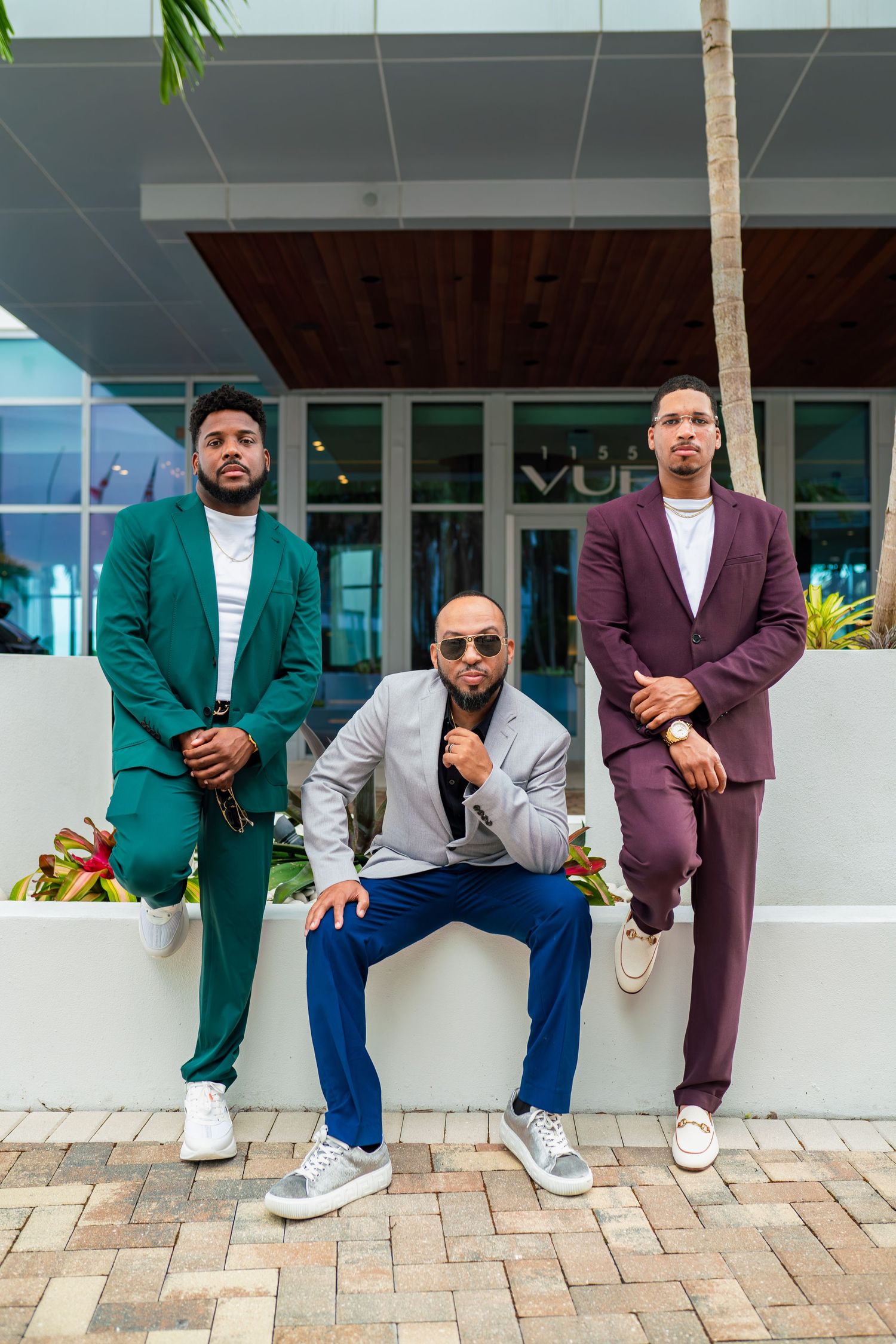 Three men in stylish suits pose against a modern building exterior with palm trees.