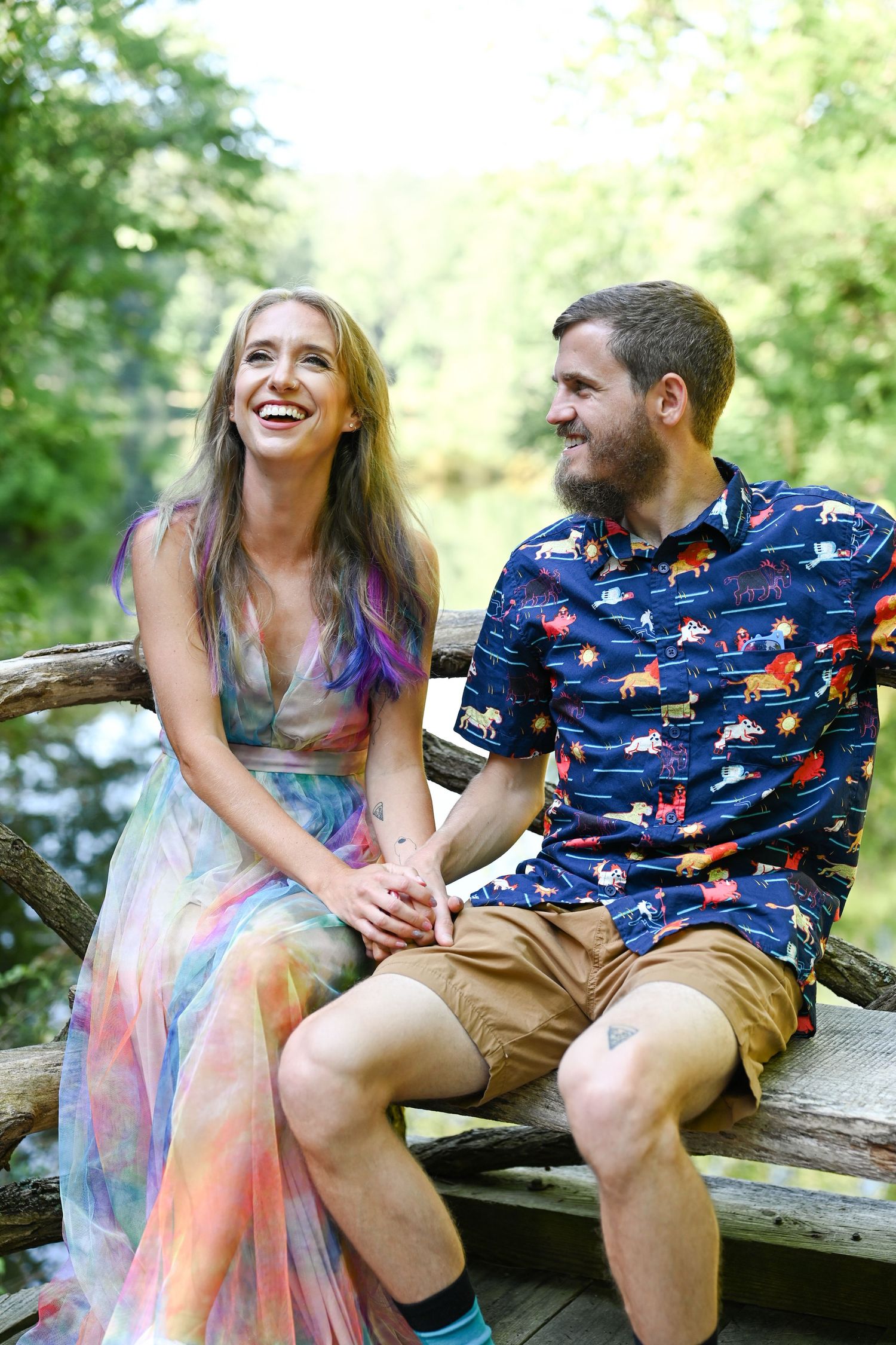 A couple laughs together while sitting on a wooden bench by a stream.