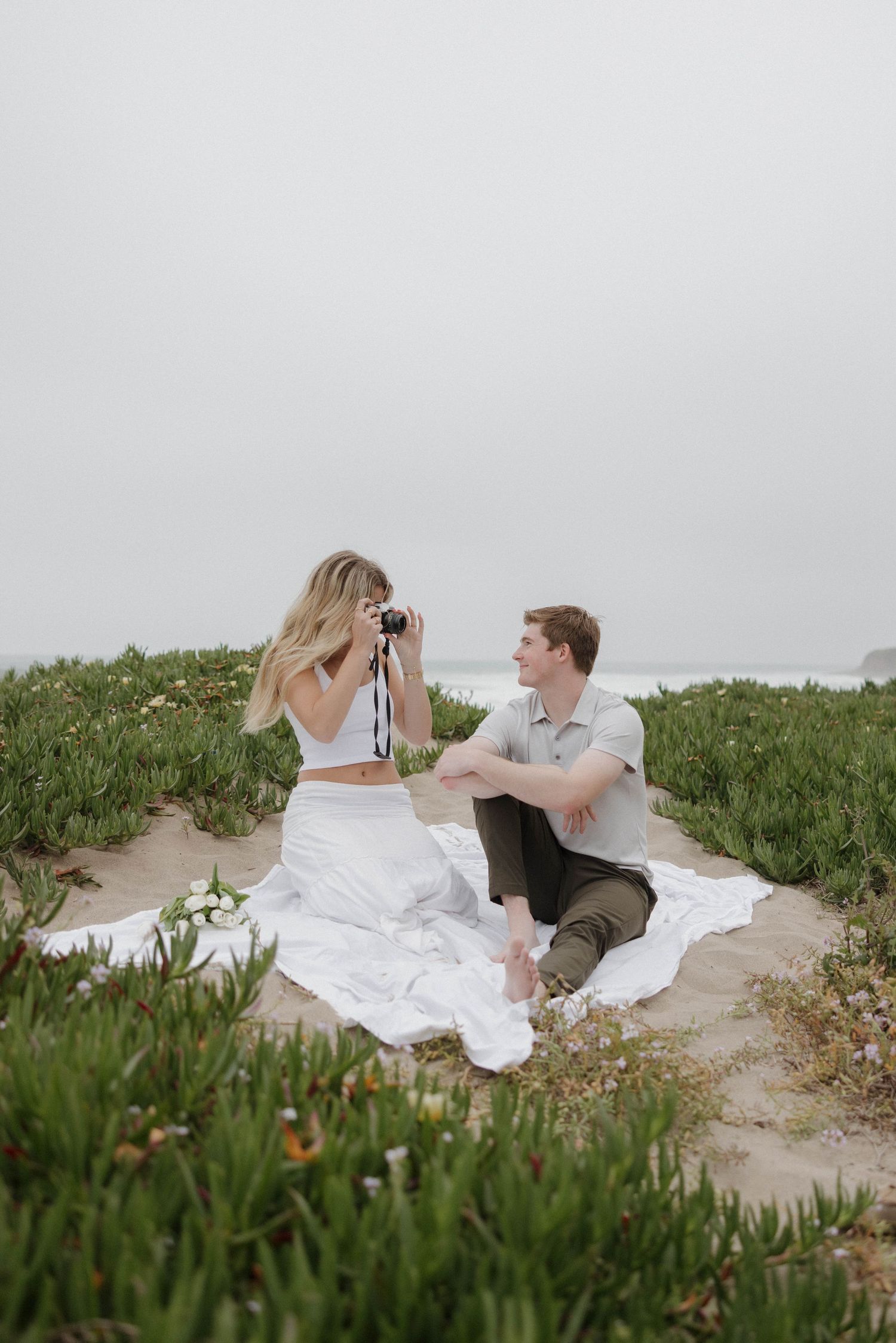 Two people sit on a white blanket on a coastal path surrounded by green vegetation on a misty day.
