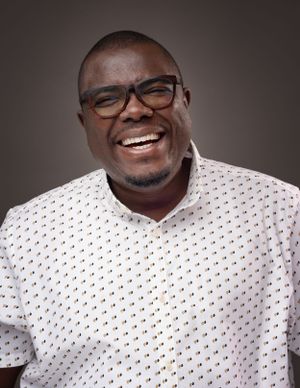 Professional headshot of a person wearing glasses and a white patterned shirt against a dark background.