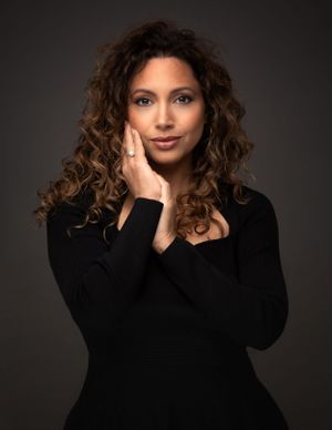 Studio portrait of a figure with curly brown hair wearing a black sweater posing against a dark background.