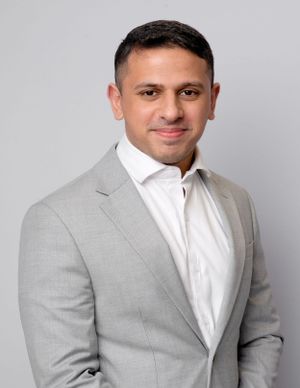 Professional headshot of person wearing light gray suit jacket and white shirt against neutral background.