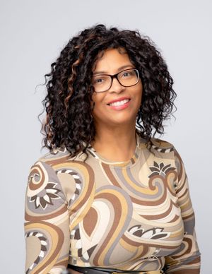 Professional portrait of a person with curly hair wearing glasses and a patterned brown top against a light background.