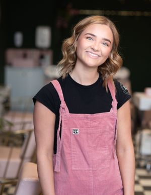 A smiling person wearing a pink corduroy overall dress with a black t-shirt underneath, posing for a casual portrait.