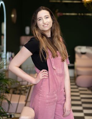 Someone posing in pink denim overalls and a black t-shirt stands in front of a checkered floor and potted plants.