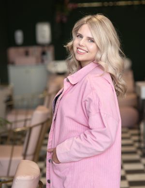 Person in pink corduroy jacket smiles warmly while standing in casual indoor setting with checkered floor.