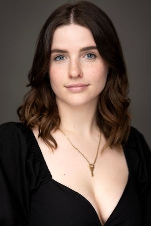 Professional headshot portrait of someone with wavy brown hair wearing a black top and delicate key necklace against dark background.