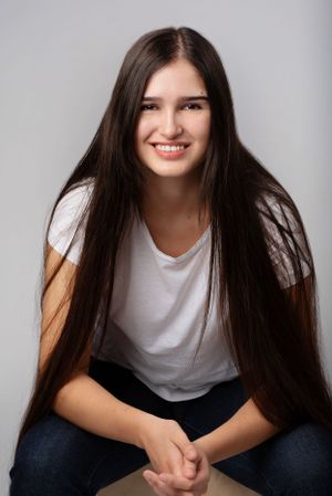 A person with long dark hair wearing a white t-shirt and jeans sits casually against a grey background.