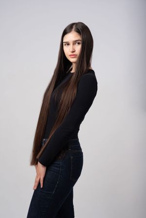 A studio portrait showing very long dark hair against a black sweater and jeans with a gray backdrop.