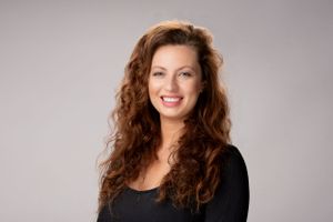 Professional studio portrait of someone with long curly brown hair wearing a black top against a gray background.
