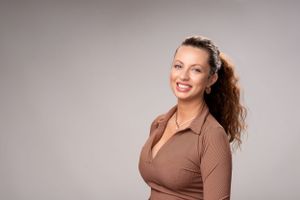Smiling woman in brown collared top with curly hair poses against gray background for professional headshot.