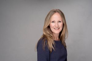 Professional headshot of someone with long blonde hair wearing a navy sweater against a gray backdrop, smiling warmly.