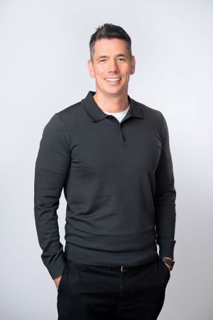 Professional business headshot of person wearing dark sweater against light gray background.