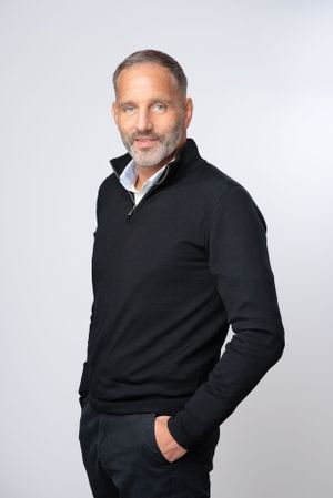 Professional studio portrait of someone wearing a black quarter-zip sweater and white dress shirt against light gray background.