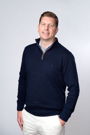 Professional studio portrait showing someone in a navy quarter-zip sweater and white pants against a light gray background.