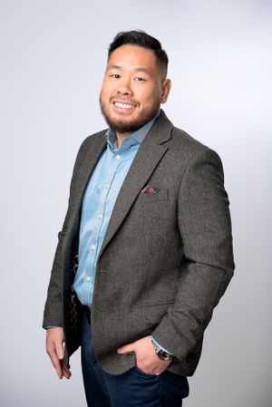 Professional headshot of a person wearing a gray blazer, light blue dress shirt and dark jeans against a light background.