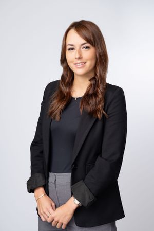 Professional headshot of someone wearing a black blazer and gray pants against a light gray background.