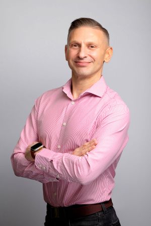 Professional studio portrait of someone wearing a pink striped dress shirt with arms crossed against a grey background.