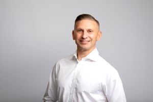Professional headshot of someone wearing a crisp white dress shirt against a light gray background.