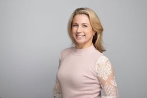 Professional headshot of person wearing a pale pink sweater with lace sleeve detail against a gray background.