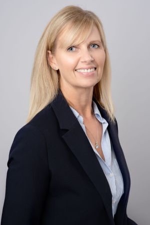Professional headshot of person in black blazer and striped button-up shirt against gray background.
