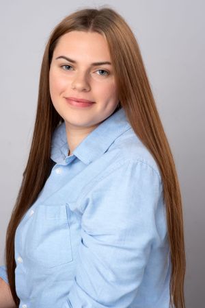 A young woman in a light blue button-down shirt smiles at the camera against a gray background.