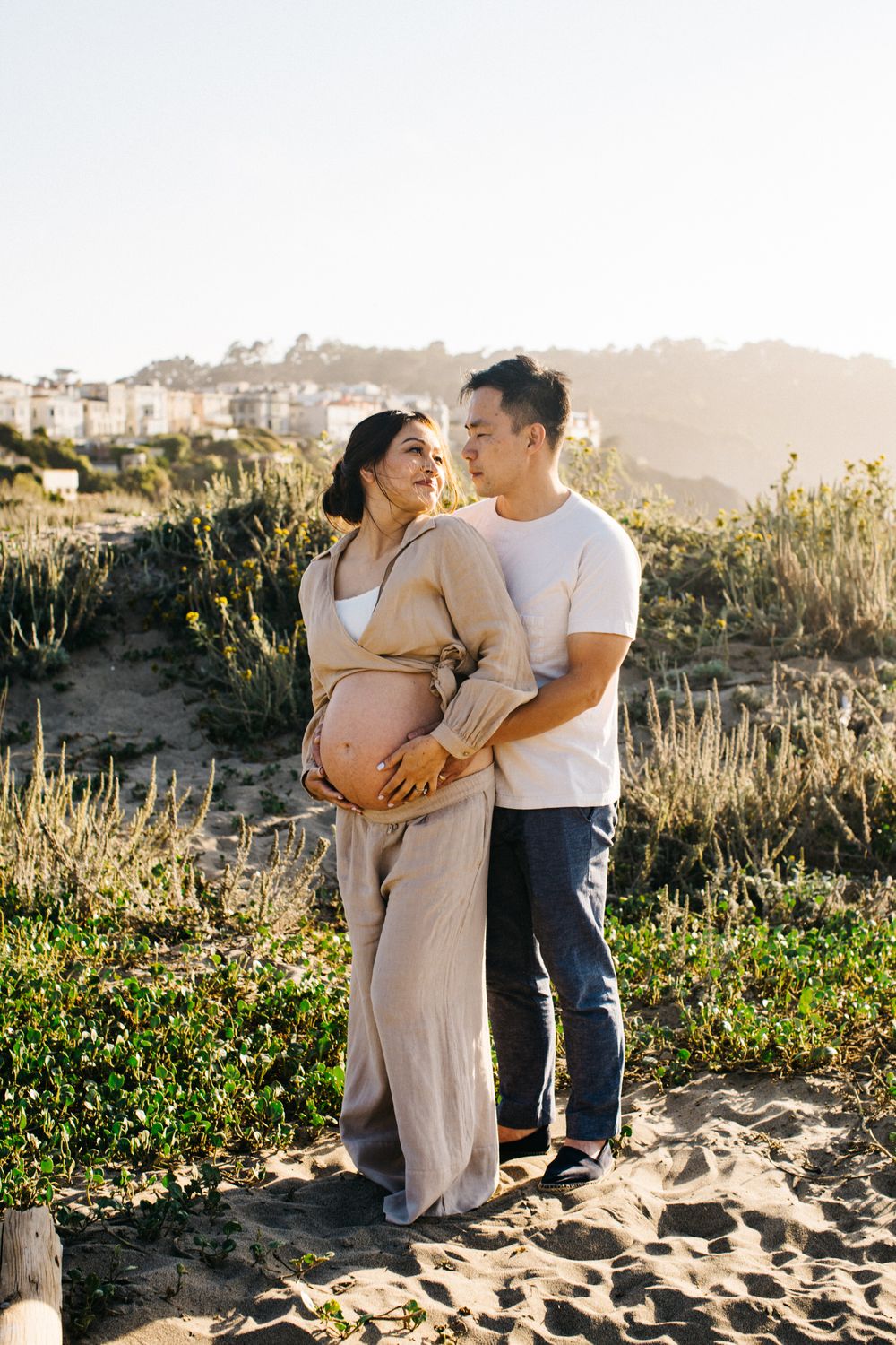 Expectant couple embracing on sandy hillside during golden hour photography session.