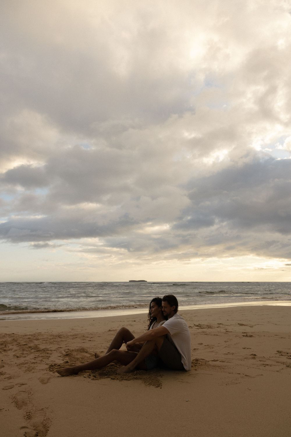 A couple sits together on a sandy beach at sunset with dramatic cloudy skies overhead.