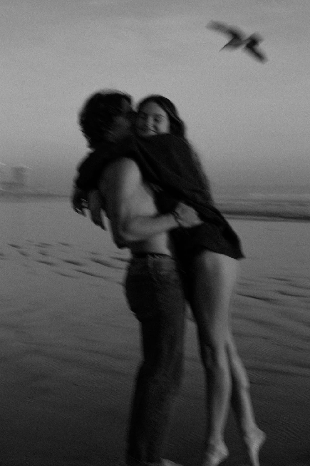 A romantic beach embrace captured in black and white with a bird soaring overhead.