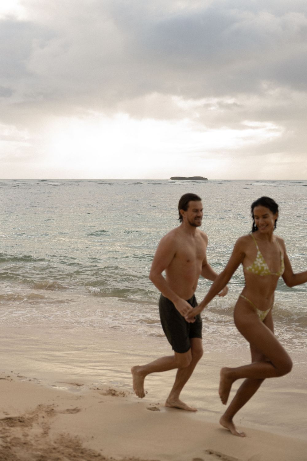 Two people joyfully running along the shoreline at dusk with waves crashing beside them.