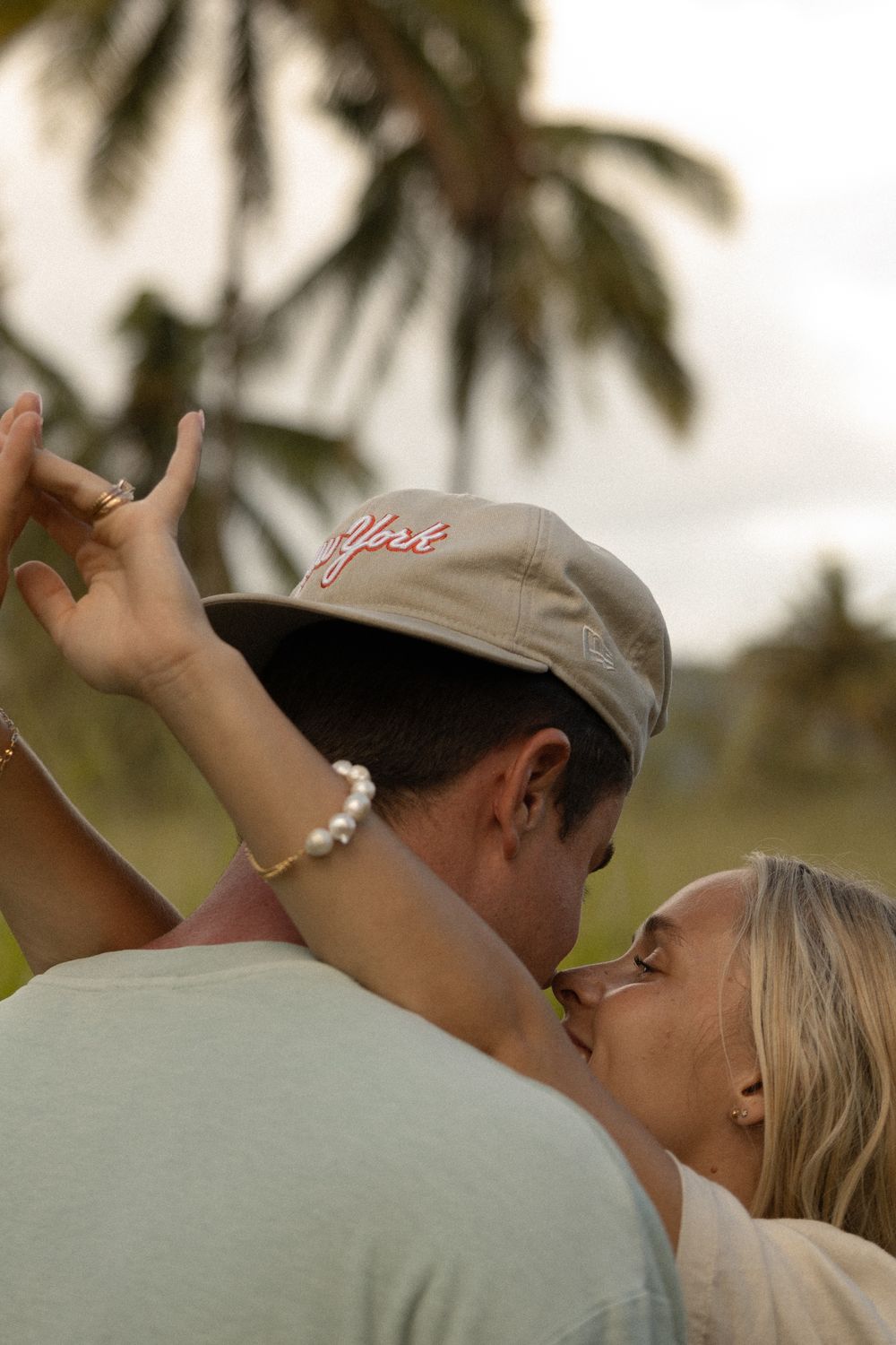 A couple shares an intimate moment under palm trees with one wearing a gray baseball cap.