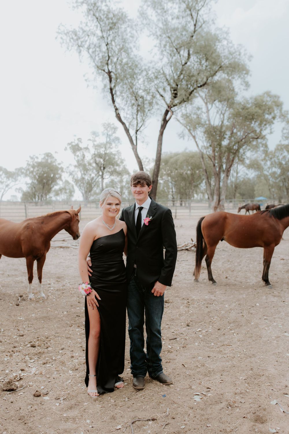 Formal couple poses with brown horses in rustic paddock setting.