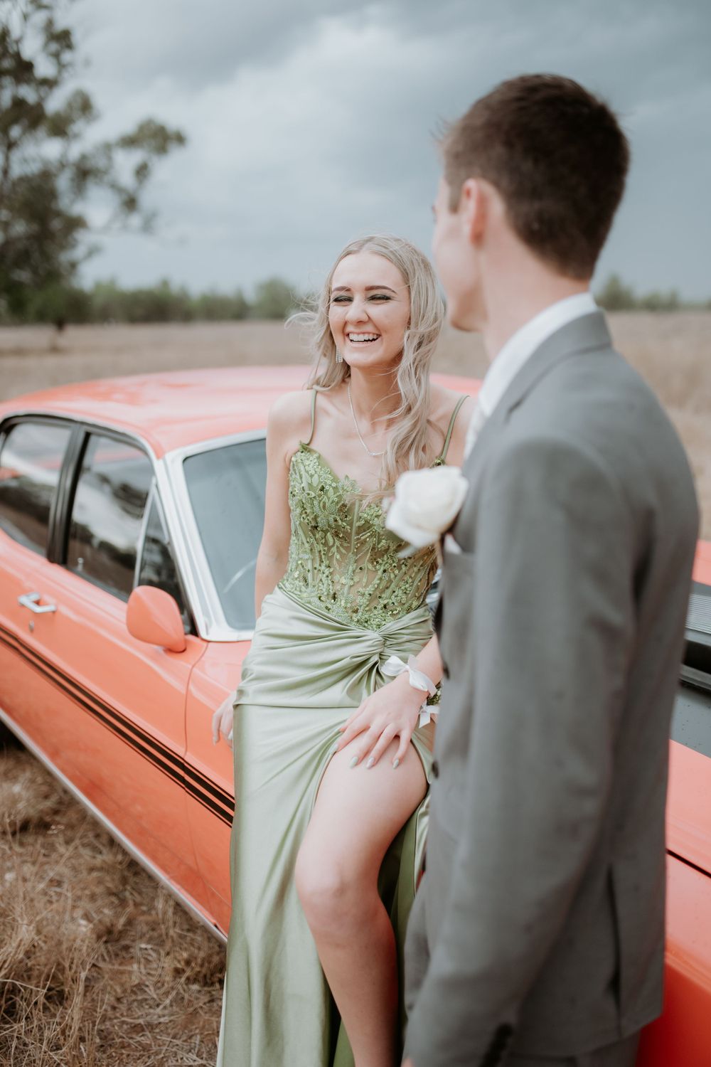 Elegant couple posing by orange vintage car in countryside setting.