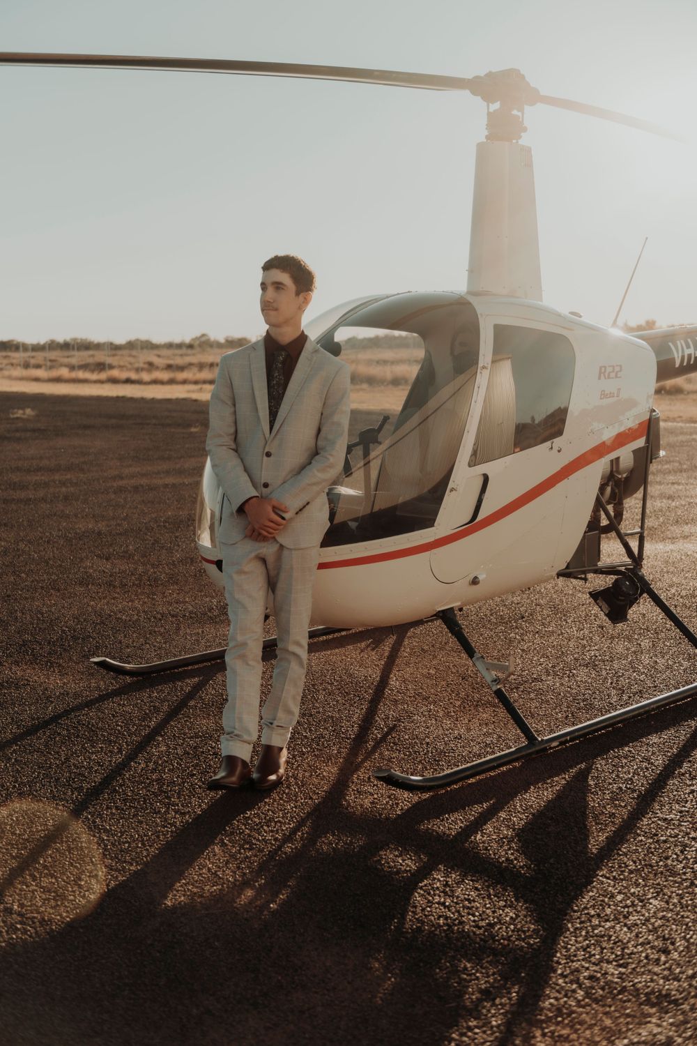 Formal portrait of someone in light suit leaning against helicopter at sunset.