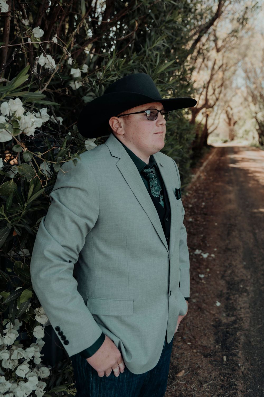 Person in gray suit jacket and black cowboy hat poses against flowering bushes.